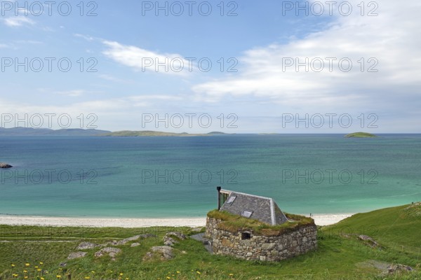 Stone cottage with grassy roof overlooking the vast sea and green hills under a blue sky, Eriksay,