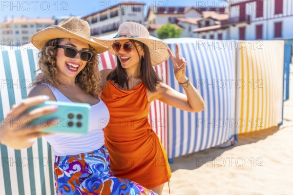 Two cheerful women wearing summer hats and sunglasses are taking a selfie with a smartphone on a