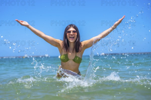 Young woman wearing sunglasses and a bikini is raising her arms and splashing water in the sea,