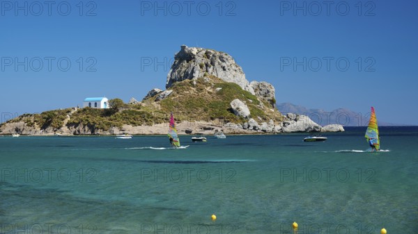 Island with windsurfers and boats in the turquoise blue sea in front of a rocky coast, Agios