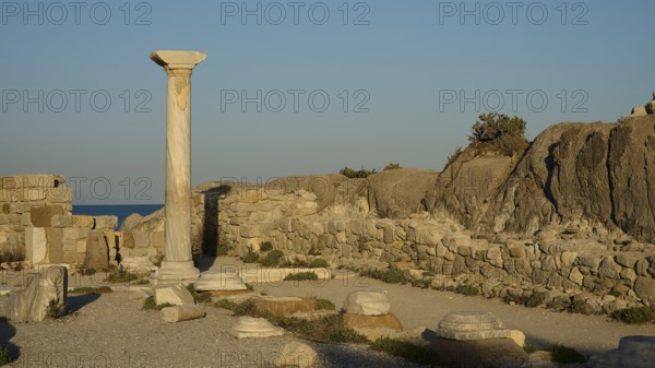 Ancient ruins with a standing stone column and surrounding rock structures in warm sunlight, Agios