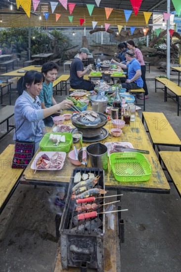 Thai man and woman cooking on a charcoal hot pot and grill at their table in a buffet style outdoor