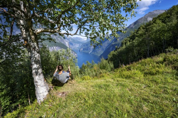 Young man swinging on a tree, view of idyllic fjord landscape of the Geirangerfjord at Blomberg