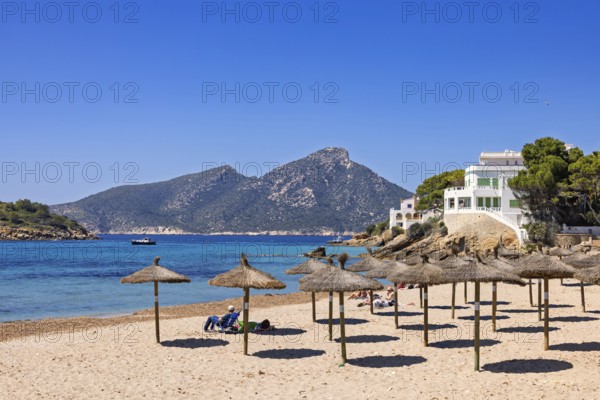 People sunbathing on a sandy beach with parasols by the Mediterranean Sea with white houses on a