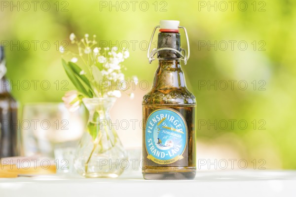 Bottle of Flensburger Strand beer on a table next to a vase of flowers in a green, natural setting,