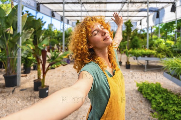 Young woman gardener with curly red hair enjoying the scent of plants, taking a selfie while