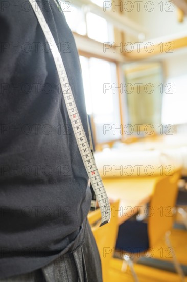 A man in a black T-shirt carries a tape measure over his shoulder in a light-coloured room with