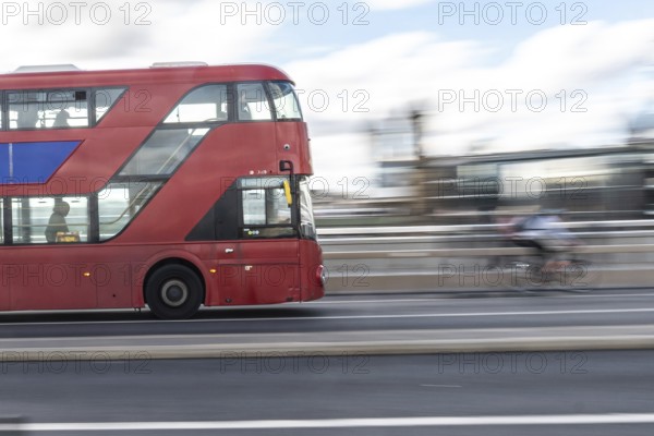 Red double decker bus driving on a london road with a cyclist passing by in the background,