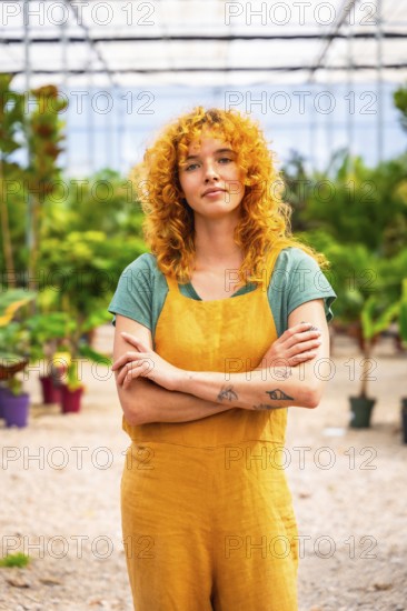 Young woman with curly red hair and tattoos wearing yellow overalls, standing with crossed arms in