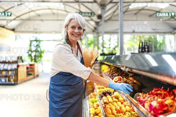 Smiling greengrocer wearing protective gloves, carefully arranging a vibrant display of fresh fruit