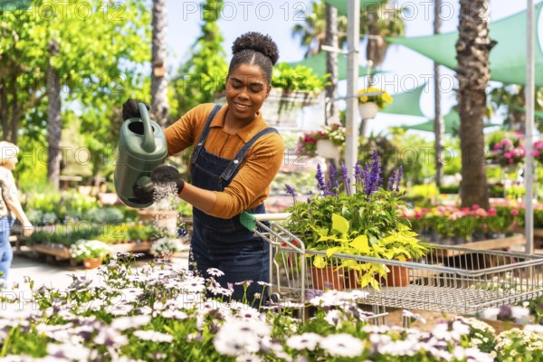 Happy young woman working in a garden center watering plants with a watering can while pushing a