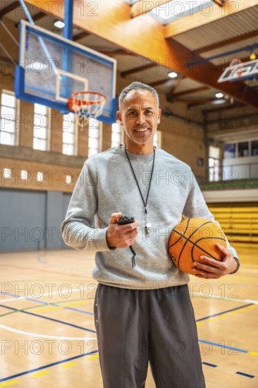 Mature basketball coach holding a ball and stopwatch, smiling confidently in an empty gymnasium,