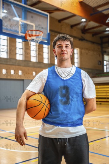 Young male basketball player holding ball in indoor court posing after training, looking confident