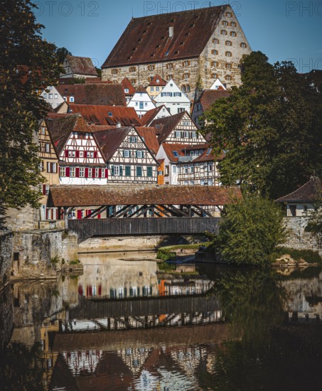 Half-timbered houses with red roofs are reflected in the river next to a wooden bridge in a
