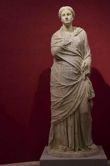 Full-length portrait of an ancient female statue in elaborate drapery against a red background,