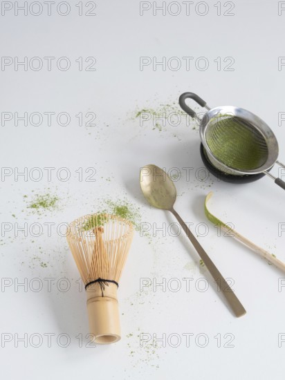 Matcha powder with traditional bamboo whisk, strainer, spoon, and scoop on a white surface