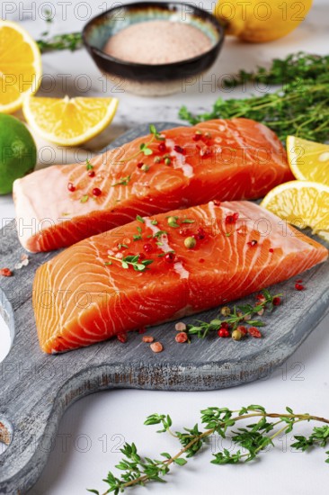 Two pieces of salmon fillet sit on a wooden cutting board. They are topped with herbs and pink