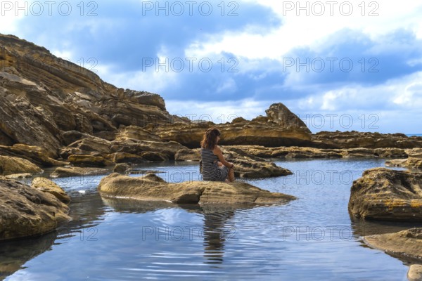 Woman sitting on a rock in a calm tide pool. Reflecting on the water. Surrounded by rugged coastal