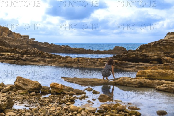 Woman crouching by a rocky tide pool on a rugged coastline, exploring the shallow water with the