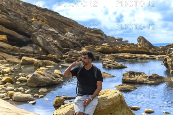 Man with a backpack sitting on a coastal rock, looking to the side and shielding his eyes from the