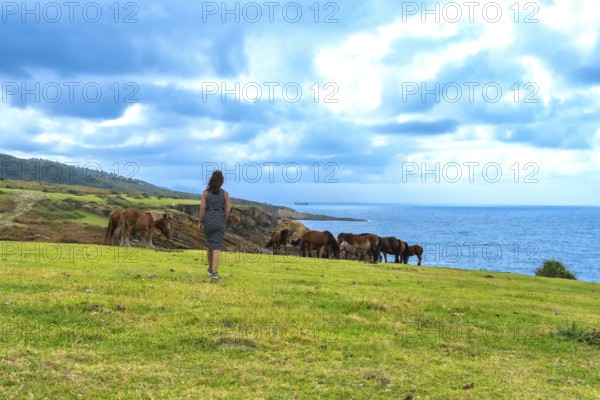 Woman standing on a green cliffside looking at a group of wild horses freely grazing near the blue