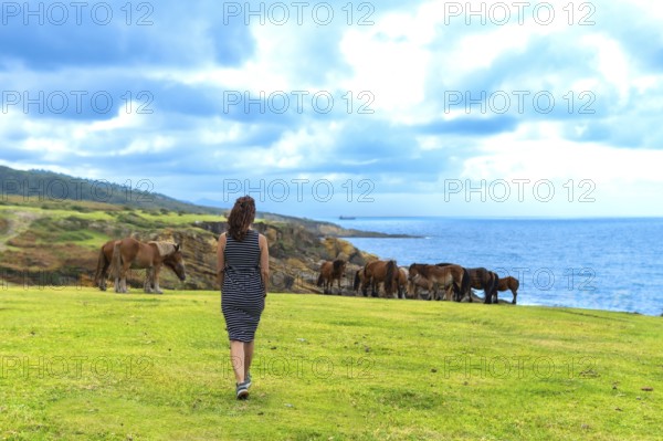 Woman walking along the verdant grass of jaizkibel mountain, observing wild horses grazing freely
