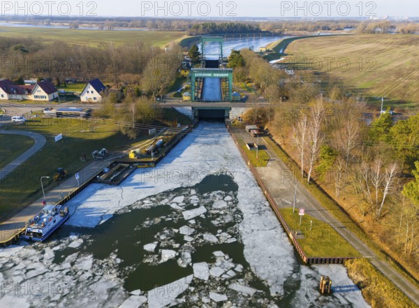 A frozen water surface in front of a lock, with gardens and trees on the edge, aerial view,
