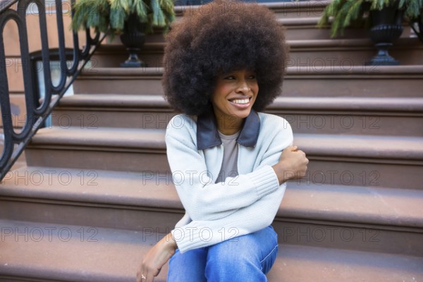 Young black woman with an afro hairstyle smiling brightly while sitting on the brownstone steps of