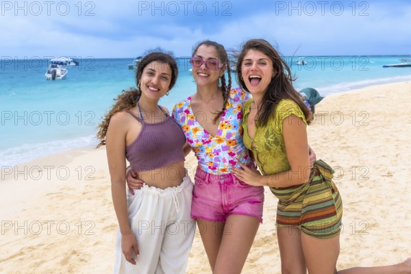 Three happy young women standing together on a white sandy beach, smiling and having fun during