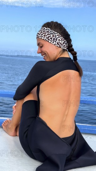 Woman wearing a wetsuit and leopard print headband, smiling happily while sitting on a boat deck,