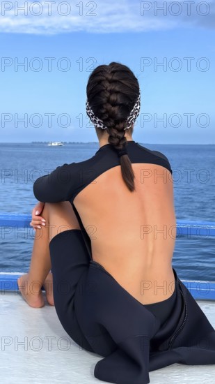 Woman wearing a wetsuit sitting on a boat deck, gazing out at the vast blue ocean and distant