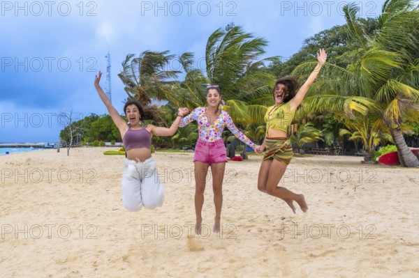 Three happy young women friends jumping while holding hands on a sandy beach, celebrating freedom