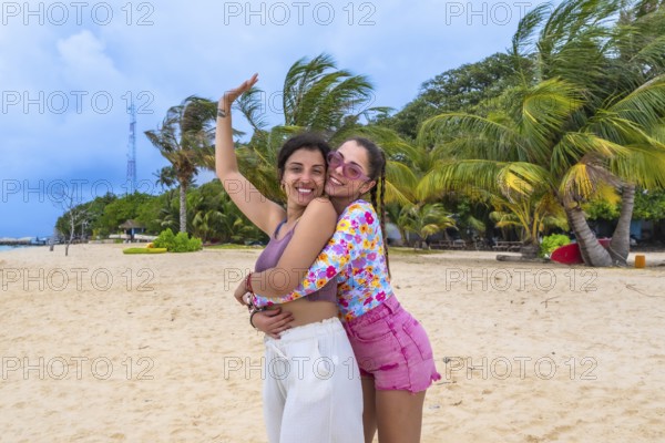 Two happy young women friends embracing on a tropical sandy beach with palm trees and a clear blue