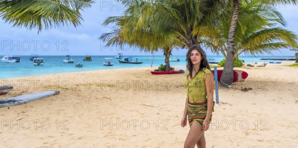 Woman stands on sunlit sandy beach under palm trees, wearing green top and sarong while gazing at