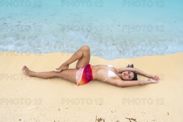 Young woman lying on the pristine sandy beach by clear turquoise ocean water, enjoying a tropical