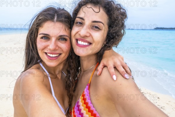 Young women smiling for a selfie on a sandy beach, embracing during a tropical island vacation,