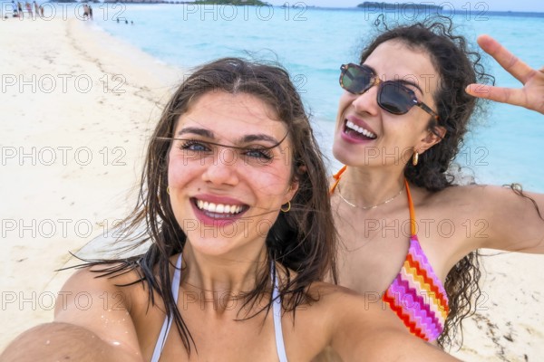 Two smiling young women taking a self portrait on a white sandy beach with turquoise ocean in the