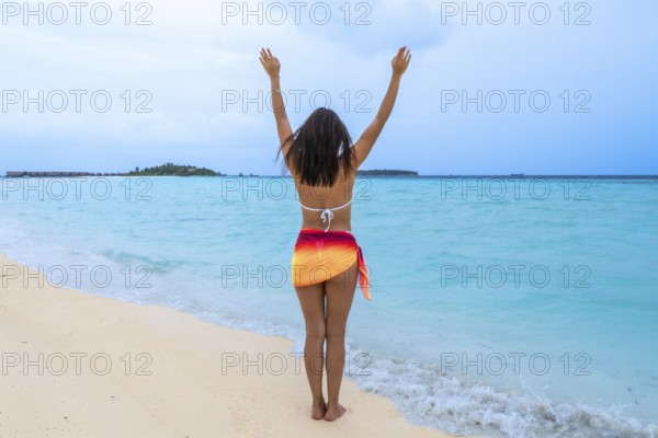 Woman standing on a white sand beach with turquoise ocean water, raising arms in joy and freedom,