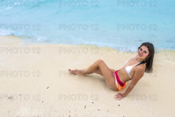 Young woman in a bikini and colorful sarong sitting on white sandy beach, smiling while relaxing by