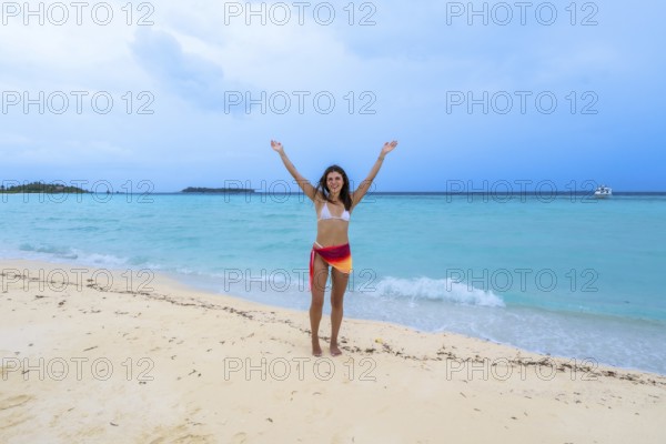Young woman standing on a pristine white sand beach with turquoise water, raising her arms in joy