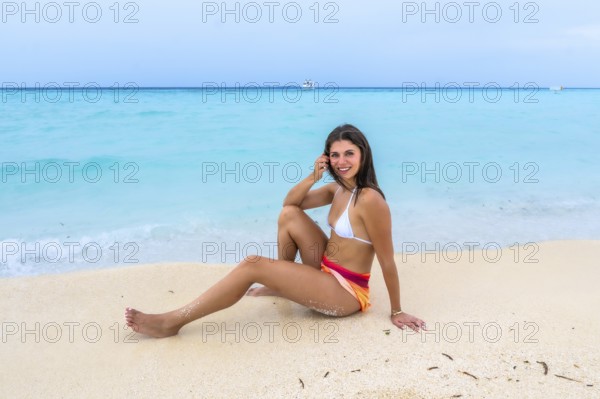 Young woman sitting on white sand beach smiling, enjoying a relaxing tropical getaway with clear