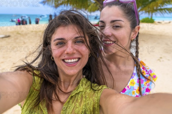 Two young happy women smiling for a close up selfie, standing on a sandy beach during a summer