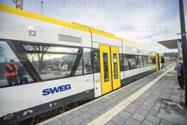 A yellow and white train is parked at a platform, passengers are waiting next to it, the Hermann
