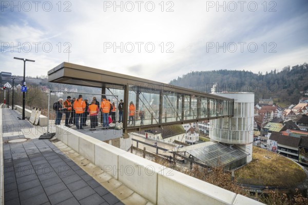 People in orange vests stand on a glass bridge with a view of a city and hills, taking off the