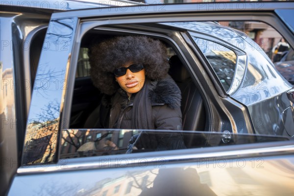 Black woman with a confident expression, wearing sunglasses and a stylish afro, is sitting in a car