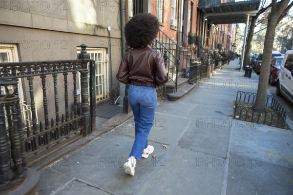 Young woman with afro hair walking along a manhattan sidewalk past brownstones, casual denim jacket
