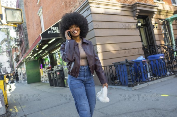 Happy young woman with an afro smiling widely, talking on her mobile phone while walking along a