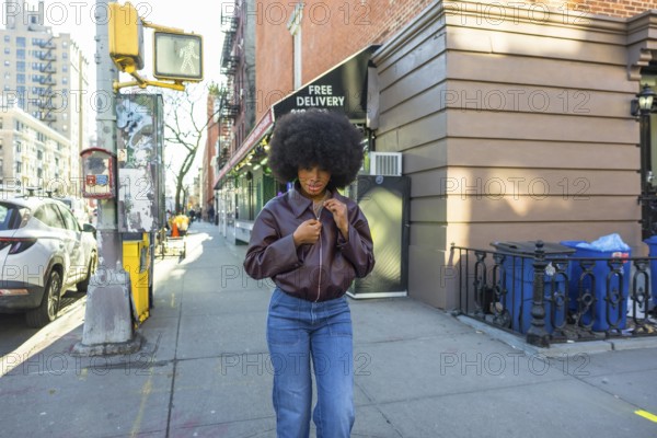 Young black woman with an afro hairstyle adjusting her brown leather jacket while standing on a