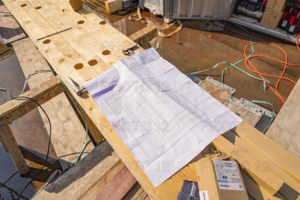 A construction plan is lying on wooden planks on a construction site, surrounded by cables and