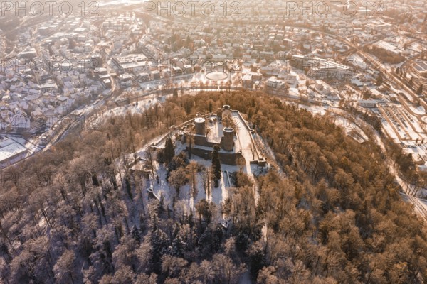 Aerial view of a snowy castle surrounded by a forest, with a city in the background at sunrise,